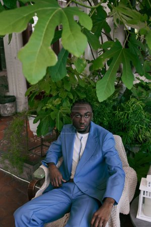 A stylish African American man in a blue suit sits on a chair in a vivid green garden.