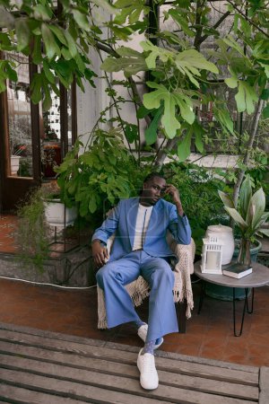 A handsome African American man in a blue suit sits on a bench in a vivid green garden.