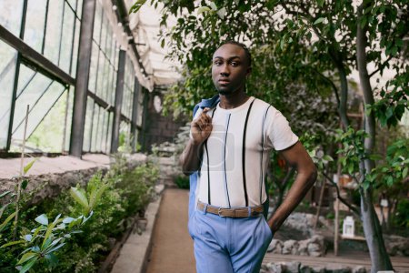 Hombre afroamericano guapo con camisa blanca sofisticada y pantalones azules posando en un jardín verde vivo.
