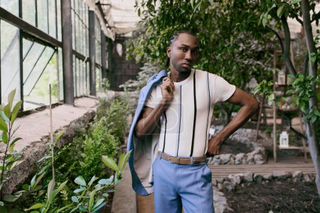 Handsome African American man in sophisticated dapper style, wearing a white shirt and blue pants, posing in a vivid green garden.