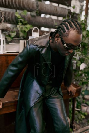 Téléchargez les photos : Un homme afro-américain élégant dans une veste en cuir noir et des lunettes de soleil pose dans un jardin verdoyant. - en image libre de droit