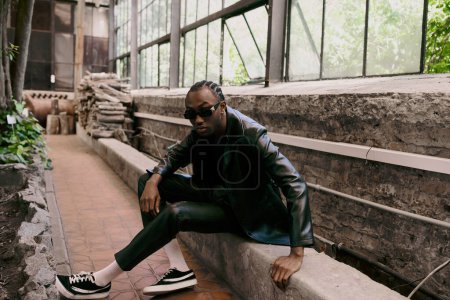 Photo for A handsome African American man with sophisticated dapper style sitting confidently on top of a stone wall in a vivid green garden. - Royalty Free Image