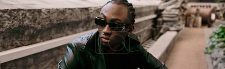 Photo for Handsome African American man in black leather jacket and sunglasses strikes a pose in a vibrant green garden. - Royalty Free Image