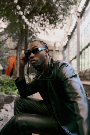 Téléchargez les photos : Un bel homme afro-américain vêtu d'une veste en cuir noir et des lunettes de soleil pose dans un jardin vert vif. - en image libre de droit