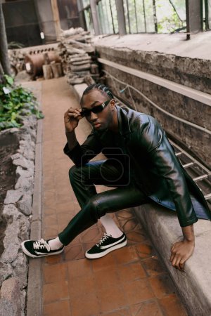 Handsome African American man in black leather jacket and sunglasses sitting on ledge.