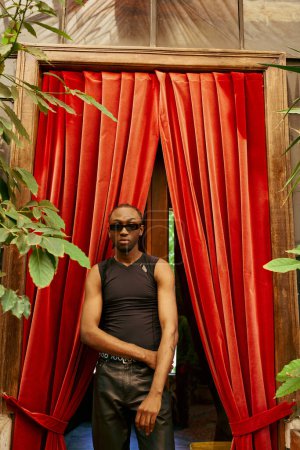 Foto de Un hombre afroamericano sofisticado se para con confianza frente a una vibrante cortina roja. - Imagen libre de derechos