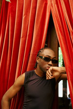 A stylish man wearing sunglasses leans against a vibrant red curtain.