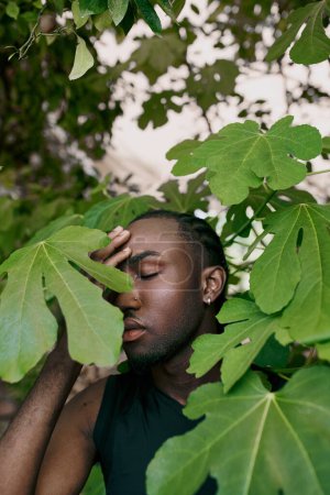 A handsome African American man with his eyes closed, hiding behind a tree in a vivid green garden.