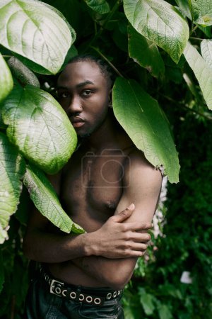 Foto de Hombre afroamericano guapo con un estilo sofisticado de pie con confianza frente a las hojas verdes vibrantes. - Imagen libre de derechos