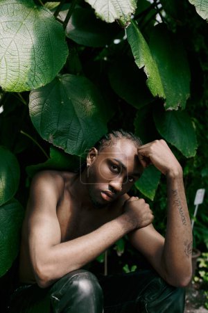 Shirtless man with dapper style relaxing by lush green leaves.