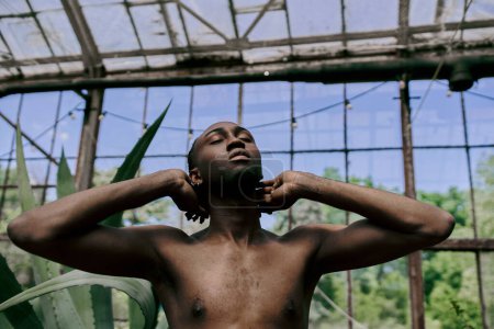 Téléchargez les photos : Un homme afro-américain élégant debout près d'une fenêtre, mains sur les oreilles. - en image libre de droit
