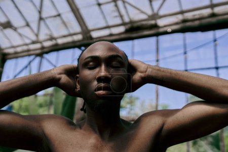 Téléchargez les photos : Bel homme afro-américain dans le style dapper, torse nu, tenant les oreilles dans un jardin vert vif. - en image libre de droit
