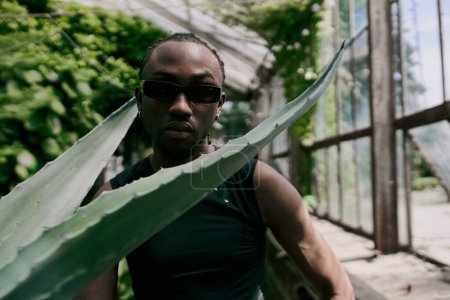 Schöner afroamerikanischer Mann mit Sonnenbrille und einem großen Blatt in einem lebhaften grünen Garten.