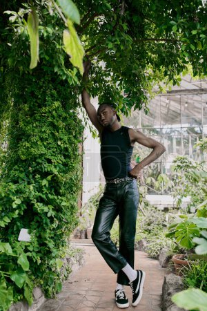 Un sofisticado hombre afroamericano vestido de negro de pie con gracia bajo un exuberante árbol verde.