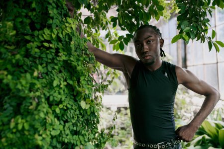 Foto de Un hombre afroamericano guapo con un estilo sofisticado se encuentra frente a una exuberante planta verde en un jardín vívido. - Imagen libre de derechos