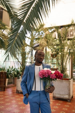 Beau Afro-Américain en costume bleu posant avec un bouquet de fleurs dans un jardin vert vif.