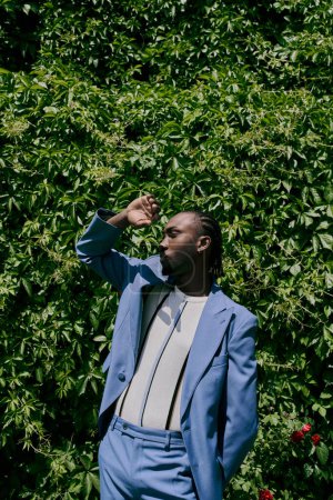 Stylish African American man in a blue suit standing elegantly in front of a lush green bush.