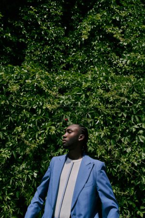 A handsome African American man in a blue suit stands confidently in front of a lush green bush.