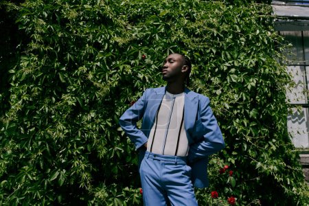 An African American man in a blue suit stands confidently in front of a lush green bush.