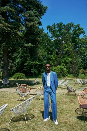 Schöner afroamerikanischer Mann im blauen Anzug inmitten von Stühlen im lebhaften grünen Garten.