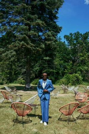 Schöner afroamerikanischer Mann im schicken blauen Anzug steht elegant in einem lebhaften Garten voller Stühle.