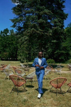 Beau homme en costume bleu sophistiqué debout dans un champ de chaises dans un jardin vert vif.