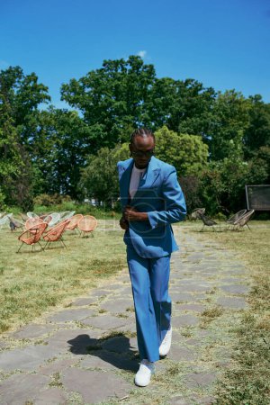 A sophisticated African American man in a blue suit walks among vibrant greenery.
