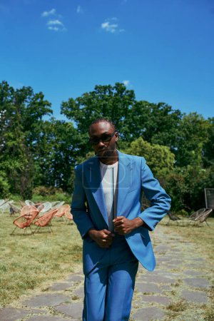 Hombre afroamericano guapo en un traje azul y gafas de sol posa en un jardín verde vivo.