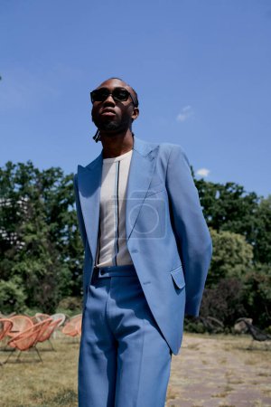Schöner afroamerikanischer Mann in blauem Anzug und Sonnenbrille posiert in einem lebhaften grünen Garten.