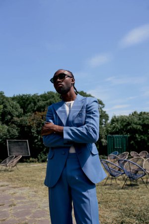 Anspruchsvoller afroamerikanischer Mann in blauem Anzug und Sonnenbrille posiert in einem grünen Garten.