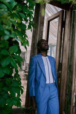 Un hombre afroamericano con un elegante traje azul se para con confianza frente a una puerta en un exuberante jardín.