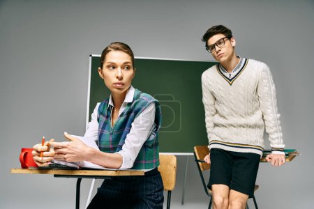 Élégant homme et femme dans un cadre universitaire, assis à un bureau devant un tableau vert.