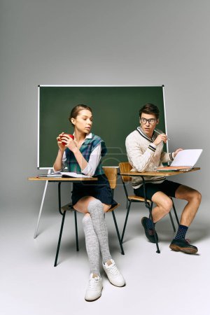 Man and woman sitting at desk, studying in front of green board.
