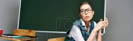 Female student in glasses sits by green board in classroom.