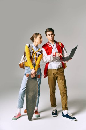 Téléchargez les photos : Un jeune homme et une jeune femme étudiants tiennent un skateboard et un ordinateur portable, debout dans une pose décontractée. - en image libre de droit