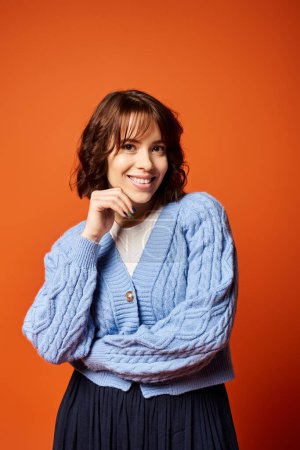 Eine junge Frau in blauer Strickjacke posiert selbstbewusst und strahlt Eleganz und Anmut in einem Studio-Setting aus.