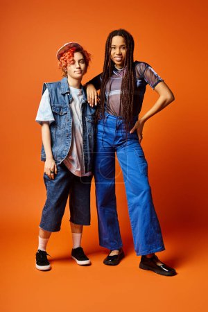 Foto de Young multicultural friends, including a nonbinary person, standing together in stylish attire on an orange background. - Imagen libre de derechos