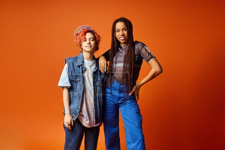 Photo for Two stylishly dressed young multicultural friends, including a nonbinary person, standing confidently next to each other in front of a bold orange backdrop. - Royalty Free Image