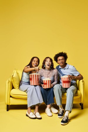 Three young multicultural friends sit on a couch, stylishly dressed, holding popcorn buckets.
