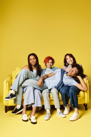 Foto de Jóvenes amigos multiculturales con un atuendo elegante sentados encima de un sofá amarillo, disfrutando de la compañía de los demás. - Imagen libre de derechos