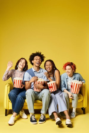 Foto de Un grupo de jóvenes amigos multiculturales, incluyendo una persona no binaria, sentados en la parte superior de un sofá amarillo vibrante en un ambiente elegante estudio. - Imagen libre de derechos
