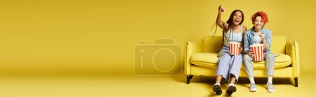 Photo for Friends in stylish attire sit on a yellow chair, happily snacking on popcorn. - Royalty Free Image