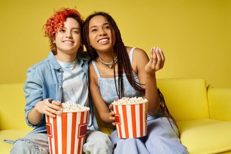 Foto de Dos jóvenes vestidos con estilo se sientan en un sofá sosteniendo cubos de palomitas de maíz, disfrutando de una noche de cine juntos. - Imagen libre de derechos