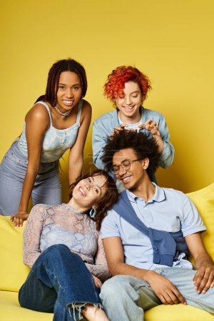 A diverse group of friends in stylish attire sitting comfortably on top of a bright yellow couch in a studio setting.