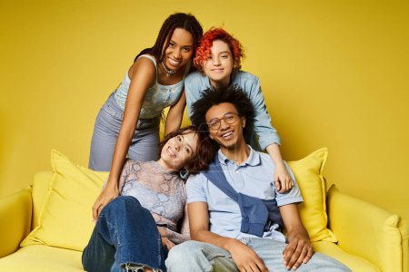 Un grupo de jóvenes amigos multiculturales, incluyendo una persona no binaria, relajándose con estilo en la parte superior de un sofá amarillo en un entorno de estudio.