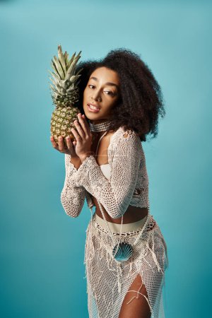 Young African American woman in trendy swimsuit holding pineapple on blue backdrop.