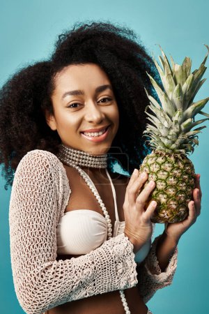 Stylish woman in swimsuit holding pineapple against blue backdrop.