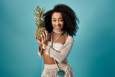 Attractive African American woman holding pineapple in trendy swimsuit against blue backdrop.