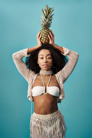 Young African American woman in stylish swimsuit balancing pineapple on head.