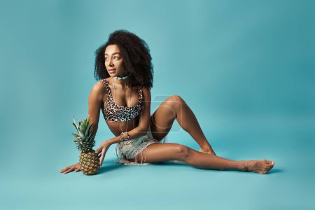Stylish African American woman in a bikini, sitting on floor with pineapple.